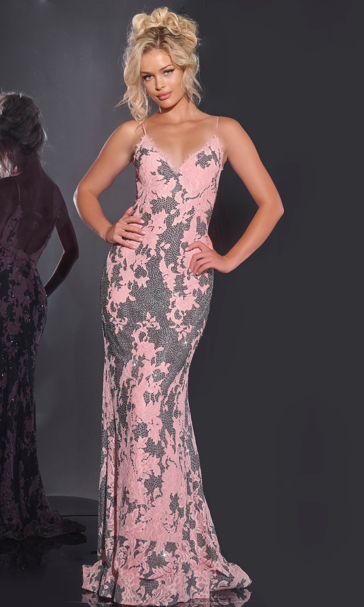 A woman models the Jovani 44033 long prom dress—a pink, floor-length, form-fitting lace gown with spaghetti straps—standing against a dark background with her reflection to the left.